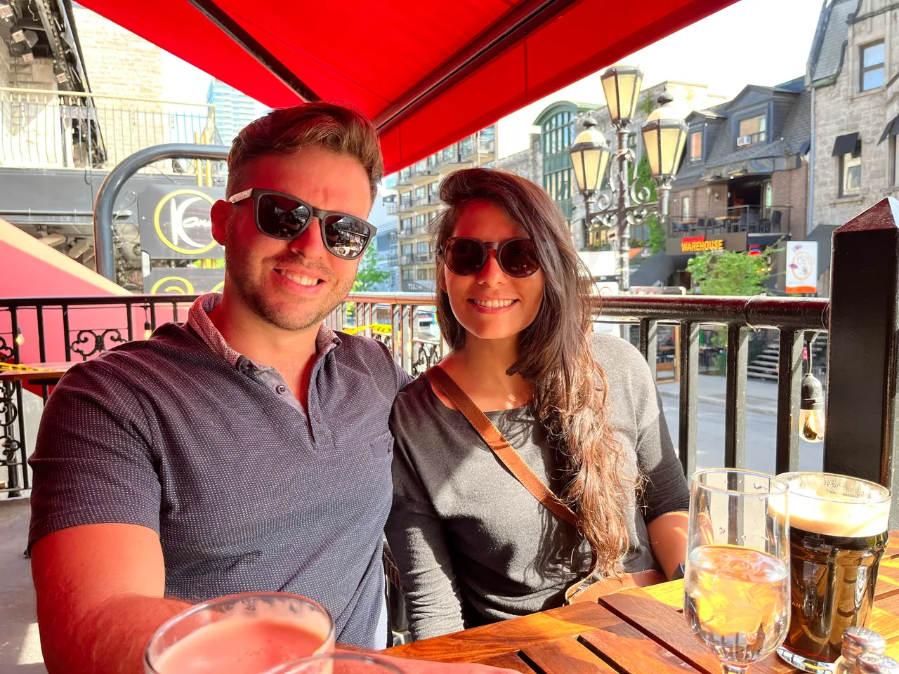 A smiling couple wearing sunglasses sits at an outdoor cafe under a vibrant red canopy, enjoying drinks.