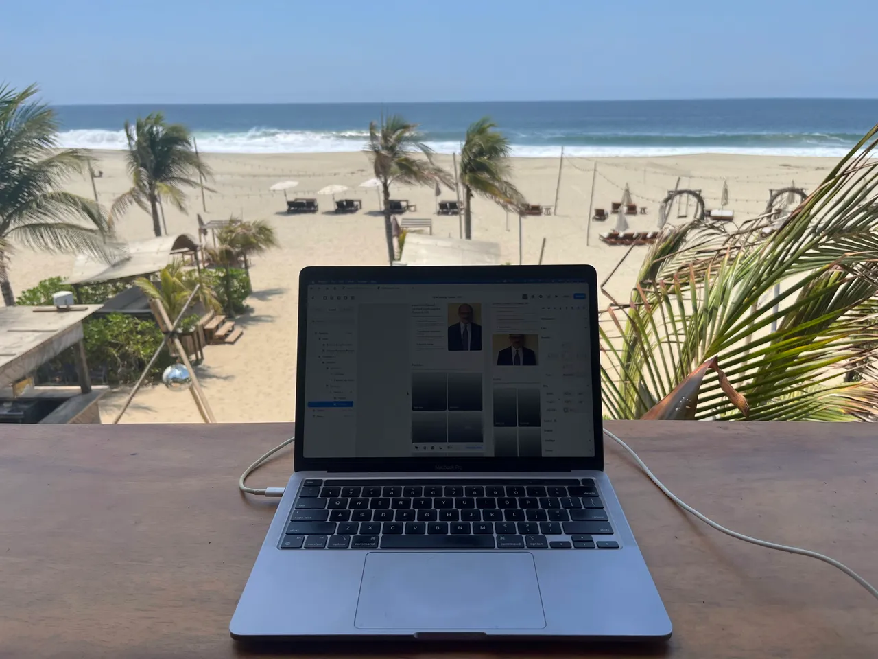 A laptop sits on a wooden desk overlooking a serene beach with palm trees.