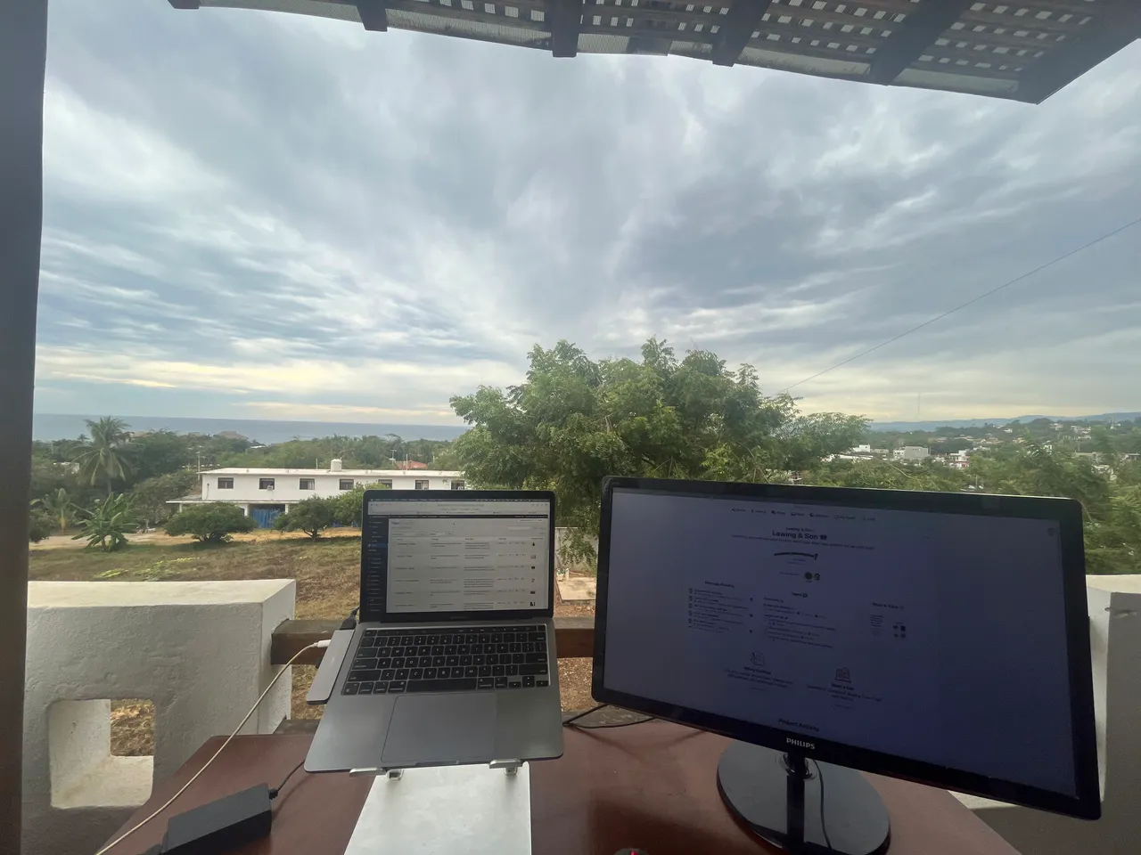 An outdoor workspace featuring a laptop and a monitor on a desk, set against a backdrop of a green landscape and a cloudy sky.