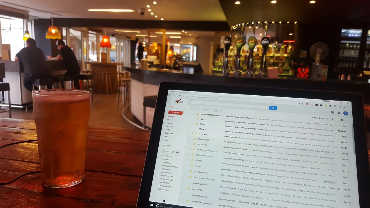 A laptop on a wooden bar table next to a glass of beer, with the pub's bar and taps in the background.